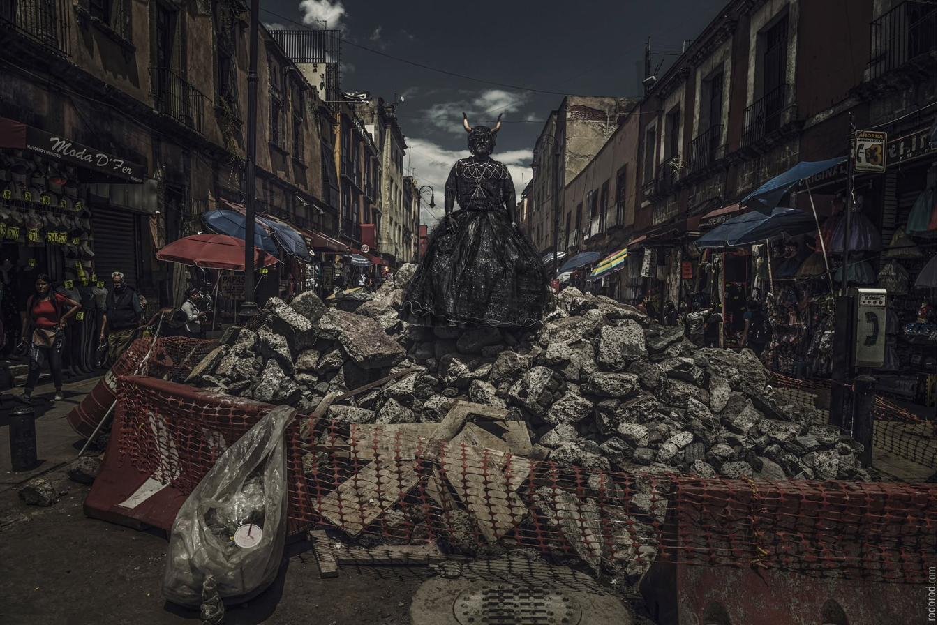 Cabaret Silencio de pie sobre un montículo de escombros en una calle del Centro Histórico de la Ciudad de México, con cuernos, collar de cadenas y falda negra.